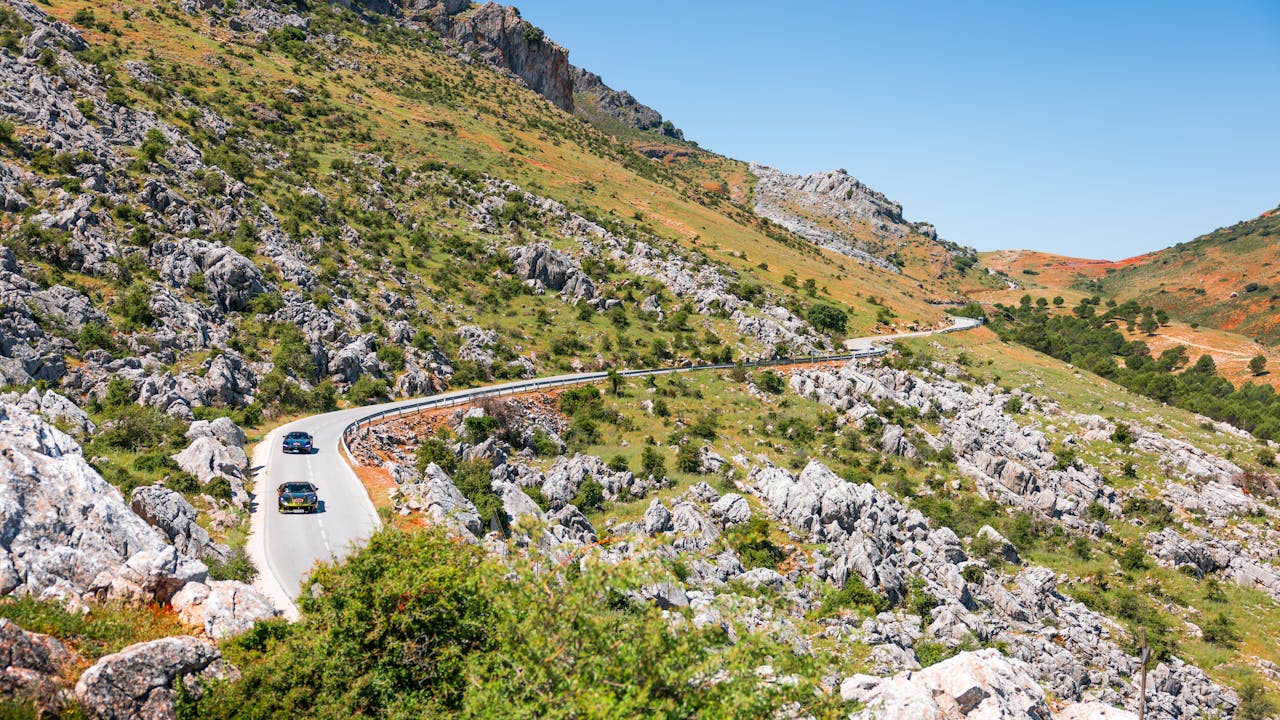 Two cars navigate a winding road through a scenic mountain landscape under clear blue skies.